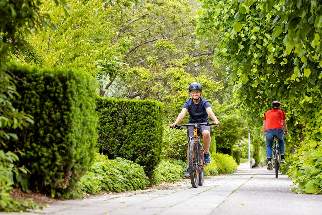 Foto på pojke som cyklar i stadsmiljö med mycket växtlighet runt omkring. En annan person cyklar åt motsatt håll i bakgrunden.