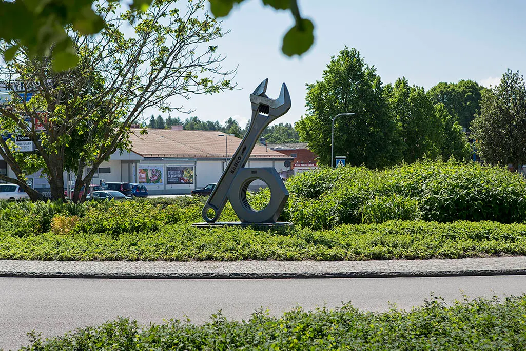 Stor skiftnyckel med mutter ståendes i en grönskande rondell med affär i bakgrunden. Foto